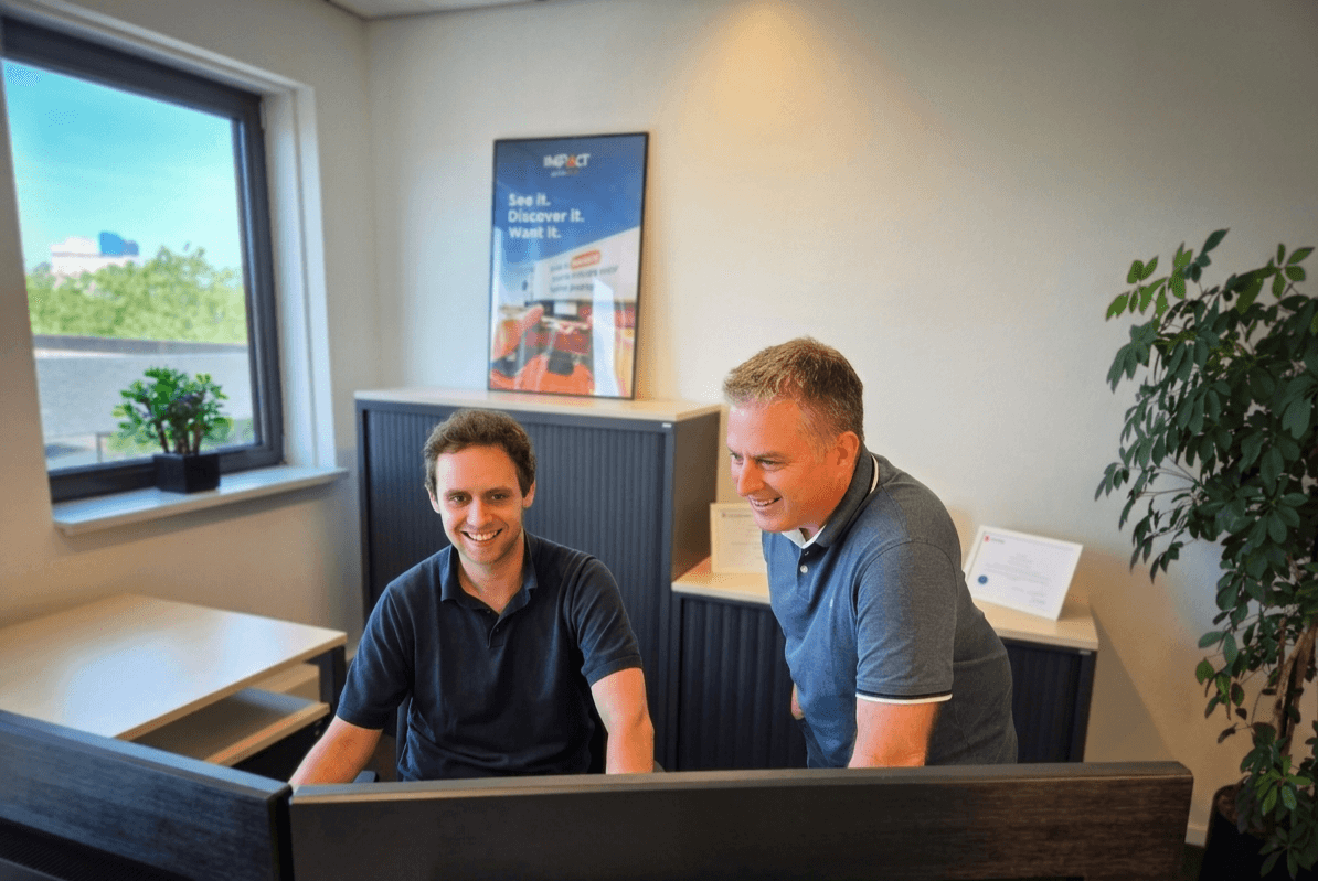 Twee supportmedewerkers achter beeldschermen in een kantoor, met een IMPACT-poster aan de muur