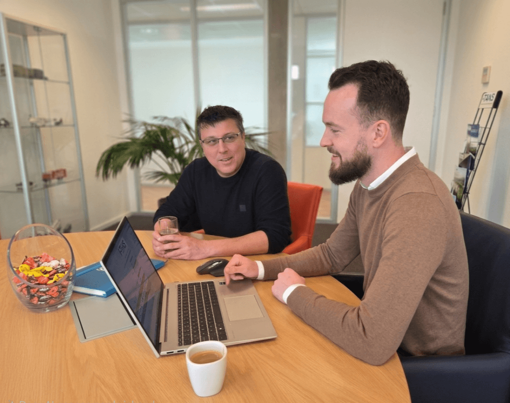 Rick en Frank van TANS overleggen lachend aan een houten tafel met laptops en koffie