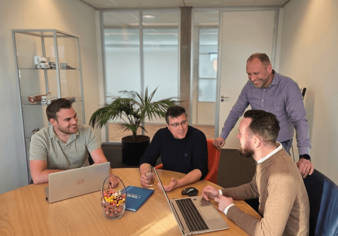 Vier TANS-collega's overleggen lachend met laptops aan een houten tafel op kantoor