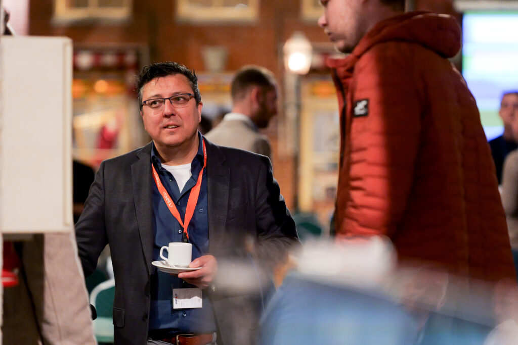 Bezoeker met bril en oranje keycord in gesprek met een kop koffie tijdens het TANS Event van 10 april