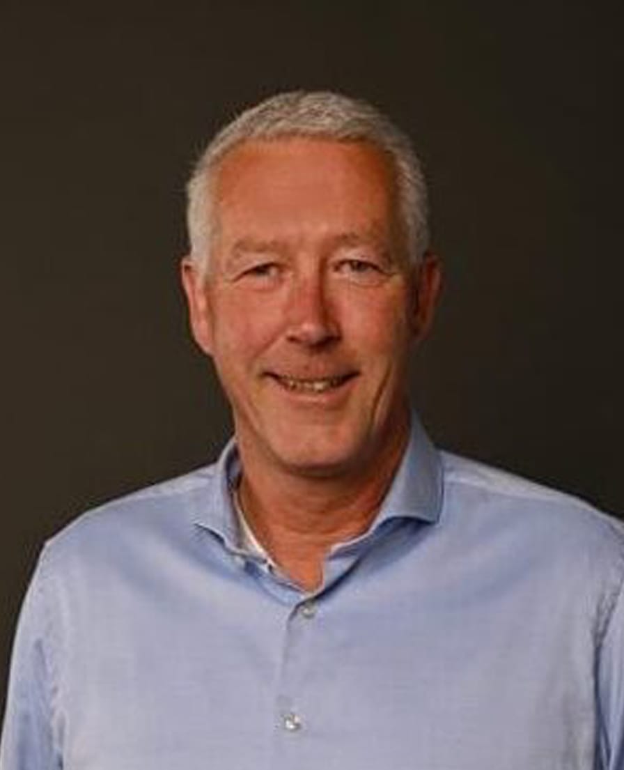 Portret van Eelko, lachende man in lichtblauw overhemd met de handen in de zakken, tegen een donkere achtergrond