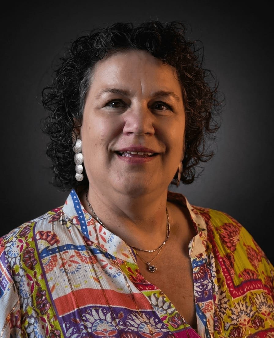 Portret van Elly met donkere krullen en een kleurrijke blouse tegen een donkere achtergrond
