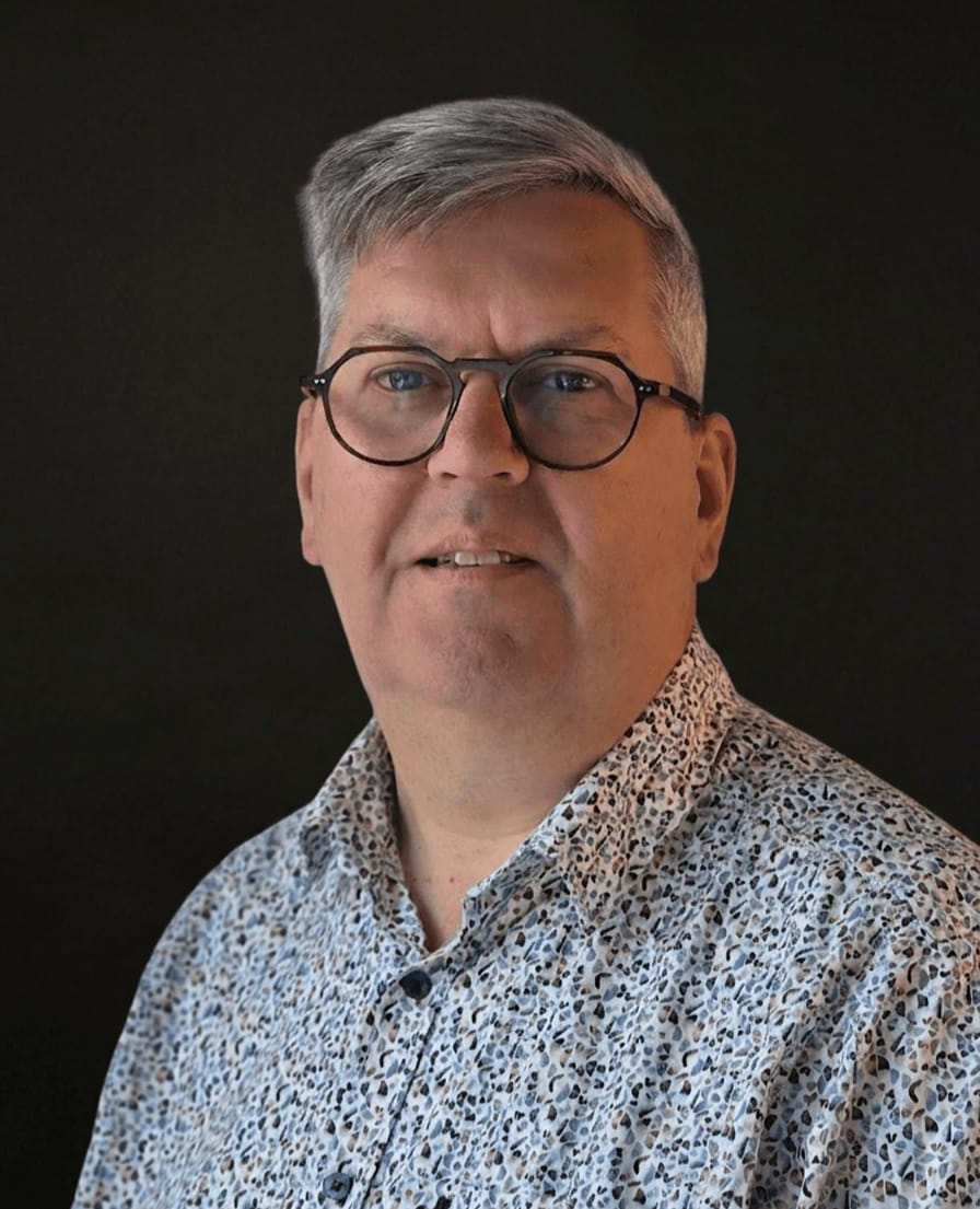 Portret van Ger met bril en lichtblauw gebloemd overhemd tegen een donkere achtergrond