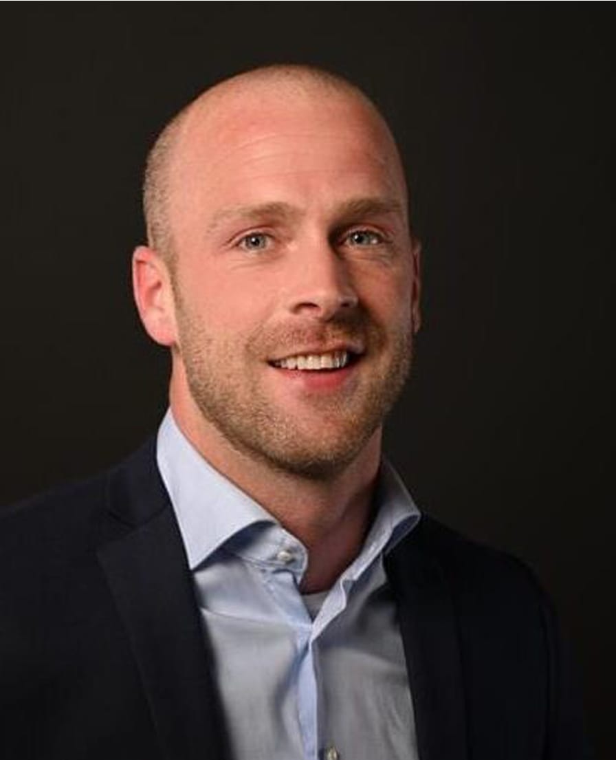 Portret van Kevin, lachend in een donker colbert met lichtblauw overhemd tegen een donkere achtergrond