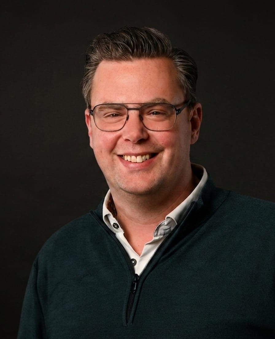 Portret van Martijn van TANS, lachend met bril en donkere trui tegen een grijze achtergrond