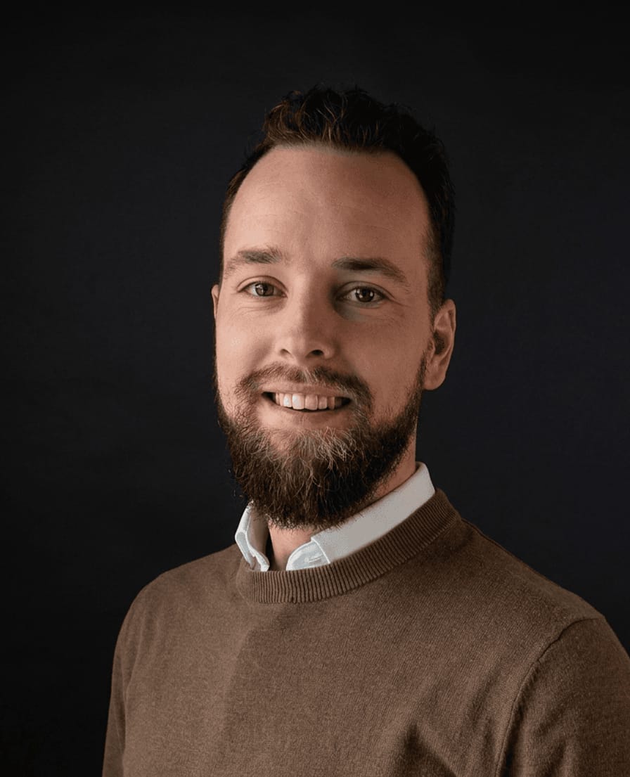 Portret van Rick, lachende man met baard in bruine trui tegen een donkere achtergrond